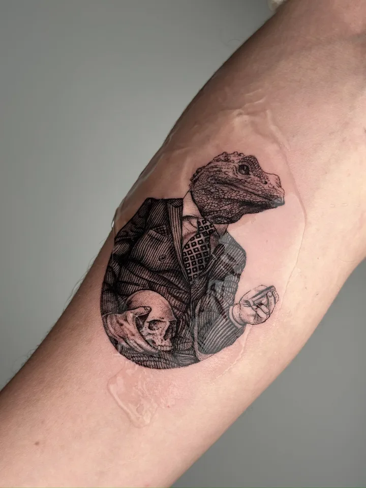 Fine-line tattoo by nurse.tattoo: a lizard in a suit holding a skull, on an upper arm