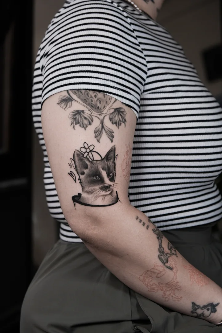 A black and grey cat portrait with a daisy behind its ear and a peony above it, on the upper arm