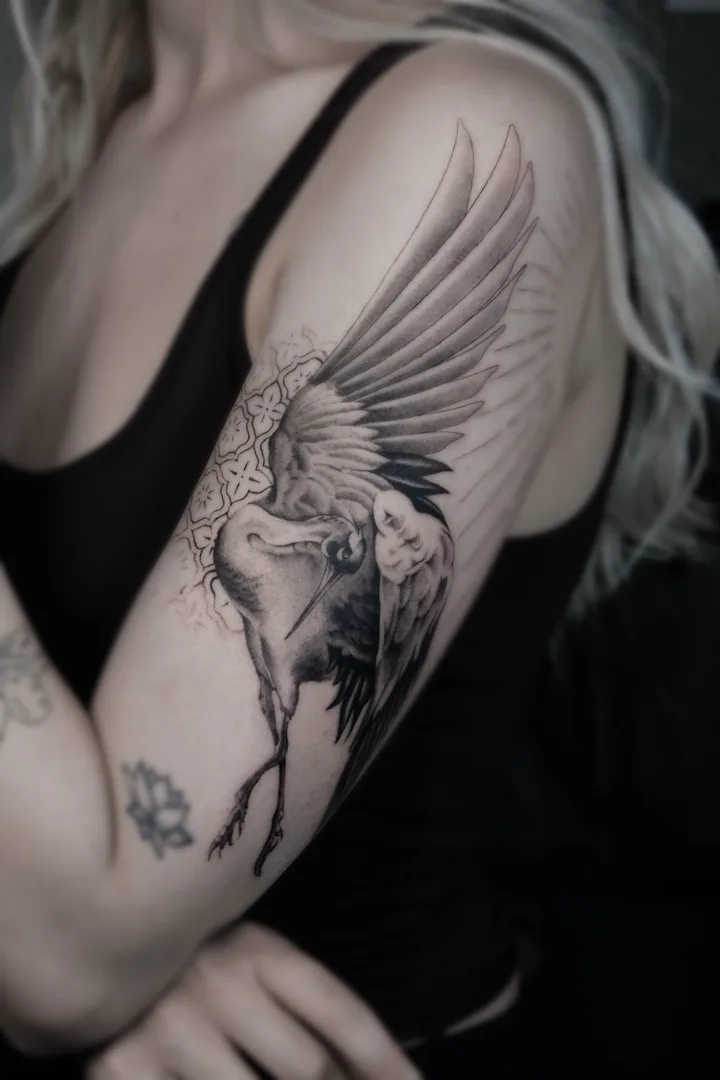 A crane in flight, wings spread over an ornamental lattice patch, black and grey on the upper arm
